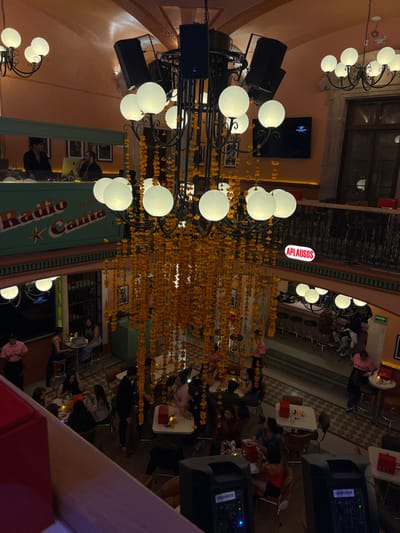 The image depicts the multi-level interior of a festive restaurant or entertainment venue located in Guanajuato City, Mexico. A dominant, large black metal chandelier with numerous spherical white light fixtures and integrated black PA speakers hangs centrally, heavily adorned with long strings of yellow-orange flowers, possibly marigolds.

On the upper level, to the left, a green booth labeled "Radio Canta" with a star logo hosts two male individuals; one appears to be operating audio equipment, while the other observes an Apple iMac. The background wall features framed pictures. To the right of the chandelier, an upper balcony with a black ornate iron railing displays a red rectangular sign with white text reading "APLAUSOS." A wall-mounted flat-screen television is visible above this railing, and another smaller chandelier with spherical lights is present.

The mid- and lower levels are filled with patrons seated at various square and round tables, engaged in dining and conversation. Several tables feature small candles or lamps. Waitstaff, identifiable by pink shirts, are present, attending to tables. A bar area with stools is visible on the mid-level right. The floor exhibits patterned tiling. In the foreground, two black PA speakers are visible, positioned below the observer's viewpoint. The venue's walls are painted a warm orange-peach tone.