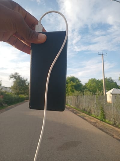 In Girei, Nigeria, a person's hand, with dark skin, holds a black rectangular power bank. A white USB cable is plugged into the power bank and curves upwards. The background features a paved road stretching into the distance, flanked by a metal fence and lush green trees. A utility pole stands tall against a sky filled with scattered white clouds.
