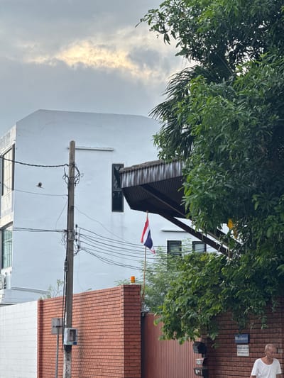 The image shows an outdoor scene in Samrong Nuea Subdistrict Municipality, Thailand. A tall white building with dark-framed windows is visible on the left. In the foreground, a brick wall runs along the right side of the frame. A utility pole with numerous wires is positioned in front of the white building and near the brick wall. A Thai flag is mounted on a flagpole near the top of the utility pole, and a small bird is in flight to the left of the pole. Dense green foliage from a large tree partially obscures the view of the white building and the sky. The sky above is overcast with a bright streak of sunlight breaking through the clouds. A man in a white t-shirt is partially visible on the far right, walking past the brick wall. The overall scene depicts a typical urban environment with a mix of modern architecture, infrastructure, and natural elements under a cloudy sky.