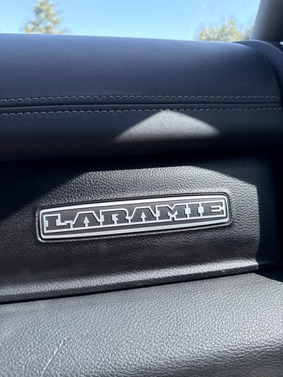 The image shows a close-up view of the "LARAMIE" emblem on the interior of a vehicle. The emblem is a rectangular badge with a silver border and the word "LARAMIE" written in raised silver letters on a black background. The emblem is inset into a textured black leather surface. Above the emblem, there is another section of black leather with visible stitching. The background is blurred, showing a bright blue sky with the tops of trees. The lighting suggests a sunny day. The scene is likely within a parked vehicle, possibly in an outdoor environment such as a parking lot or driveway in Woodland, United States of America.