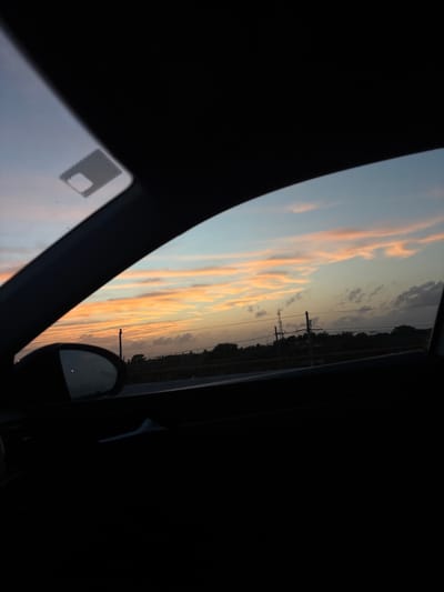 The image captures a vibrant sunset viewed from inside a vehicle. The sky is filled with streaks of orange and yellow clouds against a fading blue backdrop. Power lines and silhouetted trees run along the horizon. The car's interior, mostly in dark shadows, includes a rearview mirror reflecting the sky, a window with a sticker, and the frame of the car window, providing a partial view of the scenic sunset.