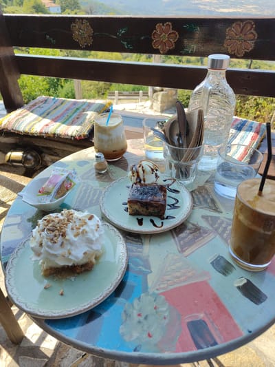 A picturesque outdoor cafe table in Palaios Panteleimonas, Greece, is set for a sweet indulgence. The table, adorned with a vibrant, hand-painted design of houses, is laden with two iced coffees, one with a blue straw and the other with a black straw. In the center, a slice of chocolate cake topped with ice cream sits on a light blue plate, drizzled with dark chocolate sauce. To the left, a portion of dessert, possibly a cheesecake, is generously topped with whipped cream and chopped nuts, served on a similar blue plate. A small white dish contains a packet of cookies. Utensils are neatly arranged in a glass, and a bottle of water stands beside it. The background reveals lush green trees and hills, hinting at a scenic mountain view. The cafe's wooden railing, decorated with floral motifs, frames the view, creating a charming and inviting atmosphere.