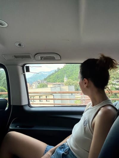A woman with her hair in a messy bun, wearing a white tank top and denim shorts, is seated inside a vehicle, looking out of the window. The view outside the window shows a town nestled against a lush, green mountainside, with buildings and trees visible. A church spire can be seen among the trees. The image appears to be taken during the daytime, with bright natural light illuminating the scene. The woman's posture and the way she is looking out suggest a moment of contemplation or observation as the vehicle moves through the landscape. The interior of the vehicle is visible, with parts of the ceiling and seats. The overall scene depicts a scenic journey through a European town. No visible text is present in the image.
