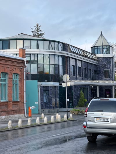 The image shows a multi-story building complex in Akhieli, Georgia. The architecture is a mix of styles, with an older brick facade featuring light blue-framed windows at the base, transitioning into a modern glass and steel structure. Above this is a section with dark blue tiled walls with rectangular windows, topped by a glass-enclosed turret.
To the right of the building is a silver Toyota Land Cruiser Prado parked on the side of the street. The license plate is visible: RSO T 115 TT. The road surface is wet, suggesting it has recently rained. The sky is overcast. Concrete bollards line the edge of the sidewalk next to the road. A street sign is visible. An orange box and a blue utility box are also present near the brick building. A small tree is growing on top of the corner between the glass section and the brick building.