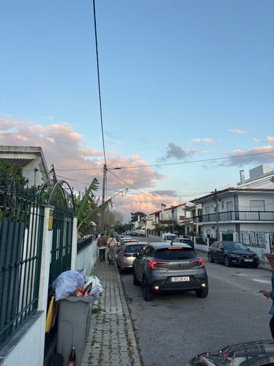 The image presents an outdoor residential street scene in Seixal, Portugal, captured under a sky displaying light blue tones with scattered cumulus clouds exhibiting pink and orange hues, indicative of late afternoon or early evening lighting.
On the left side of the frame, a dark green metal fence with an ornate arched section delineates a property. Behind this fence, green foliage, including broad banana plant leaves, is visible. Adjacent to the fence on the concrete paver sidewalk is a grey wheeled garbage bin, overfilled with a white plastic bag containing refuse. A brown glass bottle is visible on the ground directly beside the bin.
Two individuals are observed walking away from the camera on the sidewalk. The person closer to the foreground wears a light-colored jacket, patterned trousers, and a head covering. The second individual, slightly further ahead, wears a darker jacket and trousers.
The asphalt street is lined with parked vehicles. A dark grey Renault Captur SUV, bearing the Portuguese license plate "BS 26 VJ," is prominently positioned in the mid-ground, parked on the street with its brake lights illuminated. A lighter-colored hatchback is parked directly behind it. Further down the street, a dark-colored sedan with license plate "97 JZ 72" and additional vehicles are visible.
A utility pole with multiple overhead power or communication lines crosses the upper portion of the mid-ground. Residential buildings, primarily two-story structures with white or light-colored stucco facades, tiled roofs, and some featuring balconies, line both sides of the street, receding into the distance. A small portion of a person's arm, clad in a blue sleeve, is visible in the extreme bottom right corner of the image.