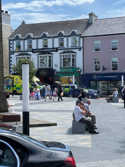 The image depicts a street scene in Donegal, Ireland. In the foreground, a person lies on a bench, partially obscured by a black car. A tall white lamppost stands prominently. The middle ground features a street with several people walking and sitting on benches. Notably, an ambulance with "PARAMEDIC UNIT" visible on its side is parked on the street. Buildings line the street, some with awnings and signage. One building has a prominent banner that reads "SELECT ACCOMMODATION". Another building displays signs for "Specsavers Opticians & Audiologists" and "the DIALCOMO CENTE". Further up the street, a building with the sign "2sk2 PHARMACY" is visible. The sky is partly cloudy, suggesting it is daytime, and the overall atmosphere appears to be relaxed, with people enjoying the outdoors. The architecture of the buildings suggests a historic Irish town.