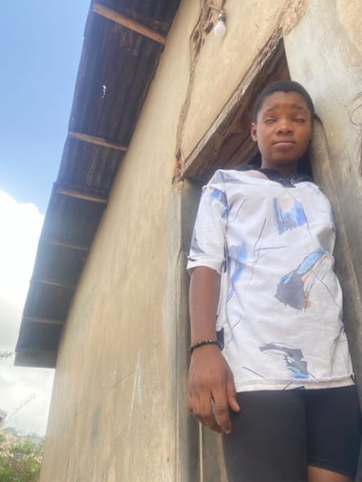 An individual, appearing female, stands leaning against a weathered wooden doorframe within a light tan-colored building in Idiroko, Nigeria. The person has dark skin, short dark hair, and their eyes are closed or appear discolored, presenting a blank facial expression. They wear a loose, short-sleeved white shirt featuring blue and grey abstract patterns, a dark collar, and black shorts or leggings. A dark beaded bracelet adorns their right wrist. Above the doorframe, a single exposed light bulb hangs. To the left, a corrugated metal roof, supported by wooden beams, exhibits discoloration and wear, beneath a bright sky with scattered white clouds. Faint greenery and distant structures are visible in the background to the left. No other individuals are present or interacting in the scene.