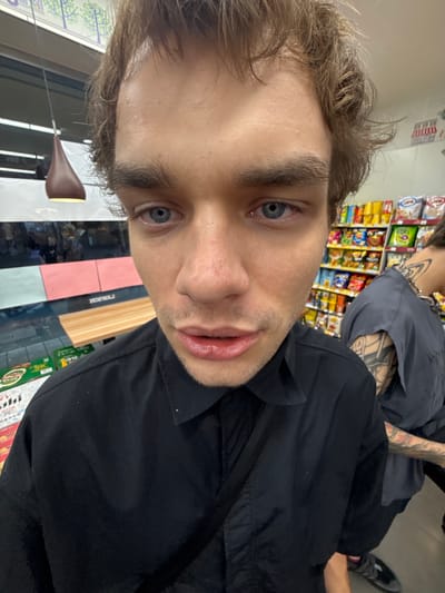 The scene depicts the interior of a convenience store, identified as a Lawson chain store by a partially visible, reversed "HOFWAS" sign on a reflective surface, located within the Changning District, China. A young male subject is featured prominently in a close-up, wide-angle perspective, facing the viewer directly.
The primary subject has light brown, wavy hair that appears somewhat disheveled and covers a portion of his forehead. His skin is fair, with visible pores and slight redness around the nose. His blue eyes are directed at the camera and appear slightly reddened. His eyebrows are thick and dark brown. The nose is prominent, showing slight redness on the tip and sides. His lips are slightly parted, revealing a fuller lower lip, and exhibit a chapped or moist texture. Minimal stubble is visible around his mouth and jawline. He is wearing a black collared button-down shirt.
To the right and slightly behind the main subject, a second individual is partially visible. This person wears a dark grey or black short-sleeved top and displays extensive black ink tattoos on their right arm and extending onto the neck and upper back, including a design resembling a collar or chain pattern around the neck. A chunky metallic chain necklace is also visible on this individual. Dark footwear is partially visible on a dark floor in the lower right corner.
The background reveals typical convenience store elements. Shelves stocked with numerous brightly colored packaged snacks and beverages line the right side of the image. On the left, a counter area features a dark surface, a wooden panel behind it, and a reflective glass surface displaying inverted reflections of people and the aforementioned "HOFWAS" sign. Below the counter, additional packaged goods, some with green labeling, are visible. A distinctive dark brown, teardrop-shaped pendant light fixture with a warm glow hangs on the left side of the scene. The overall lighting is consistent with indoor commercial illumination.