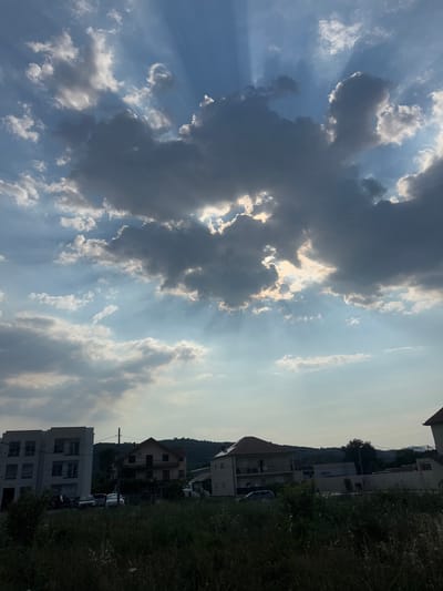 The image depicts a dramatic sky with crepuscular rays emanating from behind dark, ominous clouds, suggesting a stormy or the aftermath of a storm. The sun is partially obscured, casting a bright glow through the clouds and creating a sense of ethereal light. The sky occupies the majority of the frame, with a backdrop of a suburban or semi-rural landscape visible on the right side.
The landscape consists of several buildings, varying in architectural style and size, interspersed with dense green vegetation and trees. One building appears to be a modern, multi-story apartment complex, while others are smaller houses. A few cars are parked in front of one of the buildings. The terrain slopes upwards behind the buildings, indicating a hilly or mountainous region. The overall impression is one of a late afternoon or early evening scene, given the quality of the light and the presence of dramatic cloud formations. The location is identified as Radanovići, Montenegro. No people are visible in the image.