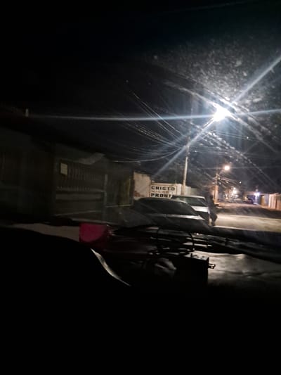 The image depicts a street scene at night in Cumana, Venezuela. A bright streetlamp illuminates a section of the road and the facade of a building on the left. The building features a barred gate and a sign with the text "CRISTO VIENE PRONTO" (Christ is coming soon). Two cars are parked or stopped on the street. The foreground is dark, obscuring details of what appears to be the interior of a vehicle. The overall impression is that of a residential street at night, with typical urban elements such as streetlights, overhead wires, and buildings.