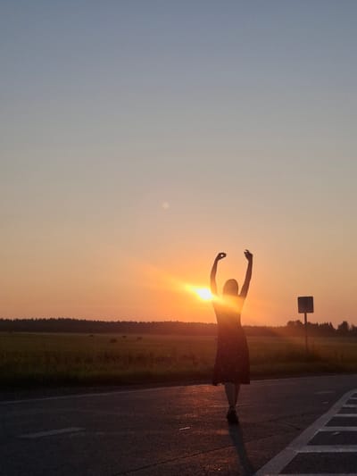 A lone individual, silhouetted against a vibrant sunset, stands on a paved road with their arms raised. The individual wears a dark, patterned dress and appears to be walking away from the viewer. The setting is rural, with a field of hay bales visible in the background, bordered by a line of trees. A solitary signpost stands to the right of the individual. The sky transitions from a pale blue at the top to shades of orange and yellow as it nears the horizon, where the sun is setting. The scene is captured in the twilight of day.