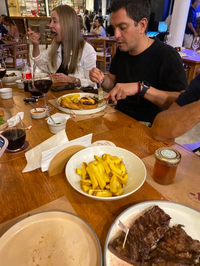 The image shows a table inside a restaurant in Punta Hermosa, Peru, located at the coordinates provided (latitude: -12.333, longitude: -76.826). The table is made of wood and is set for a meal with dishes of food, glasses of wine, and napkins. Two people are seated at the table. A woman, wearing a white shirt and dark pants, is sitting on the left side of the image, and she is looking to the right. A man, wearing a black shirt and black pants, is sitting on the right side of the table. He is looking towards the left of the image, using his right hand to hold a knife and his left hand to hold a fork. The man is cutting a burger on a plate that is in front of him. There are various food items, such as french fries and steak, present on the table. There are also glasses of red wine and a bottle of Coca-Cola. The environment is brightly lit, and the restaurant appears to be upscale and modern.