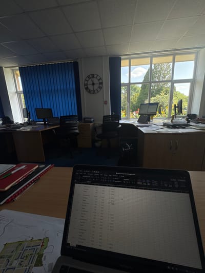 The image displays the interior of an unoccupied office space located in Mamhilad, United Kingdom. The room features a grid-patterned ceiling and blue carpeting. Natural light illuminates the scene, entering through large windows, one of which is partially covered by blue vertical blinds, while another provides an unobstructed view of outdoor trees and flowering plants.
In the immediate foreground, a silver MacBook Pro laptop is open on a light-colored wooden desk, displaying a spreadsheet application. To the left of the laptop are stacked notebooks, one red and one black, with two pens resting on top. A printed map or site plan is partially visible beneath these items.
Beyond the foreground desk, multiple workstations are arranged. To the mid-left, a darker wooden desk with a black office chair, two computer monitors, and a keyboard is positioned in front of a window fitted with blue vertical blinds. Further to the right, a lighter-colored desk is visible, featuring two computer monitors, an office chair, and scattered papers. A light-colored two-door cabinet is situated beneath this desk.
A large black circular wall clock with Roman numerals, indicating approximately 11:55, is mounted on the far wall between the two windowed sections. Below the clock, a small red square object, likely a fire alarm call point, is visible on the wall. Several filing cabinets are distributed throughout the office space. No individuals are present, and no actions or interactions are depicted within the scene.