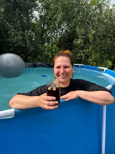 A smiling woman with reddish-brown hair tied up in a bun, wearing a wet black t-shirt, is in an above-ground swimming pool. She is holding a glass of dark liquid with ice cubes. The pool is blue with a white rim and a white pole visible on the right. In the background, there is a large grey exercise ball floating in the pool and trees. The location is specified as the city of Хохряки, Russia.