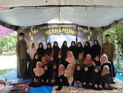 A group portrait of twenty-two individuals, comprising two adult males and twenty female children or youth, is captured under a temporary white canopy tent in Jambo Dalam, Indonesia. The individuals are positioned facing forward, with the males standing on either side of the group and the females arranged in three rows, some standing, some kneeling on a patterned rug laid over a blue tarp.
The two adult males wear patterned long-sleeve shirts, one with a black peci (cap) and dark sarong, the other with a dark peci and dark trousers. The female children and youth are dressed in Islamic attire, consisting of long-sleeved tops or dresses and headscarves (hijabs), predominantly in dark colors such as black or dark brown, with several individuals wearing lighter-colored hijabs in shades of beige, light brown, or light pink.
Behind the group, a black banner is displayed, featuring text in Indonesian "LEMBAGA PENDIDIKAN ISLAM YARHAMUNA" (Islamic Education Institution Yarhamuna), accompanied by crescent moon and hanging lantern motifs in gold and white. The tent structure includes visible white support poles and overhead wires. On the blue tarp to the far left, a pink plastic plate, a small red container, and dark cables are visible. The outdoor environment surrounding the tent features lush green foliage and trees under bright daylight. A white object, possibly a motorcycle, is partially visible in the far-right background beyond the tent.
