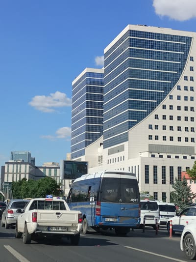 The image depicts a street scene in Yenimahalle, Turkey, on a sunny day with a clear blue sky and scattered clouds. The foreground features a traffic jam with several vehicles. A white pickup truck with license plate "06 FBA 709" is prominent, followed by a silver sedan. A blue and white passenger van with license plate "06 J 1486" is also visible. In the background, modern high-rise buildings dominate the skyline. The architecture of these buildings is characterized by glass facades and geometric designs, with one building featuring a distinctive curved white exterior. Trees and smaller buildings are interspersed between the larger structures. The overall impression is one of urban development and a busy city environment.