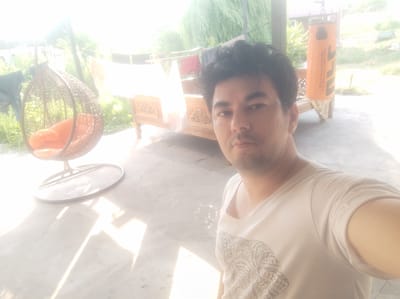 In an outdoor setting, likely a patio or veranda, a man with dark hair and a light-colored t-shirt with a printed design of an elephant is taking a selfie. He is positioned in the foreground, facing the camera. Behind him, to the left, is a hanging rattan egg chair with an orange cushion. Further back, a wooden bench with intricate carvings is visible, and above it, laundry hangs on a clothesline. To the right, an orange punching bag with black lettering hangs from a support. The ground appears to be concrete, and sunlight casts strong shadows, suggesting it is daytime. The background shows greenery and a structure with a reddish roof, possibly a house, indicating a semi-rural or suburban environment. The image is overexposed in some areas due to bright sunlight. The location is identified as Mingchinar (Blagoveshchensky), Uzbekistan.