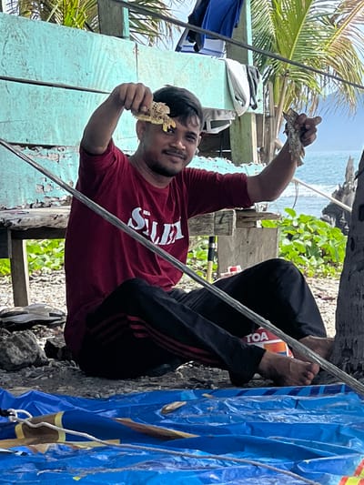 In Babahie, Indonesia, a man sits on the ground in front of a rustic, light blue wooden structure, likely near the coast. He holds up two small, patterned fish he appears to have caught. The man wears a maroon t-shirt with white lettering and black pants with red stripes. Behind him, palm fronds and the blue ocean are visible, suggesting a tropical, coastal environment. A blue tarp lies in the foreground, on which a fishing net and other fishing equipment are placed. The scene depicts a moment of successful fishing by a local resident.