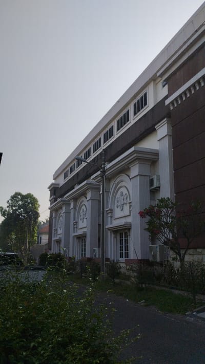 The image displays the exterior of a modern building with classical architectural elements. The building has a light-colored facade featuring prominent columns and arched decorative motifs above windows. The upper section of the building is darker, with a band of evenly spaced dark windows. A streetlamp is visible in front of the building. The surroundings include greenery, with trees and bushes in the foreground and to the left. The sky is clear and light. A paved path or road runs alongside the building. The architecture suggests it could be a public building, institution, or office complex. The presence of air conditioning units on the facade indicates a warm climate. The image captures the building from a low angle, emphasizing its scale.