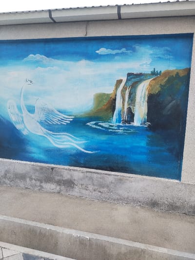 A mural is depicted on a textured grey wall in Chighirik, Uzbekistan. The artwork features a vibrant blue sky with scattered white clouds, transitioning into a vast expanse of blue water. On the right, a series of multi-tiered waterfalls cascade from brown and dark earth-toned cliffs into the water below, creating white foam and ripples. Green foliage is visible atop the cliffs. On the left, a large white swan-like bird with extended wings and long, flowing tail feathers appears to be soaring or emerging from the blue water. The mural is framed at the top by the continuation of the grey wall and a white roof gutter system. Below the mural, a concrete structure composed of grey ledges or steps, exhibiting signs of wear and minor discoloration, occupies the foreground. No individuals are present in the scene.