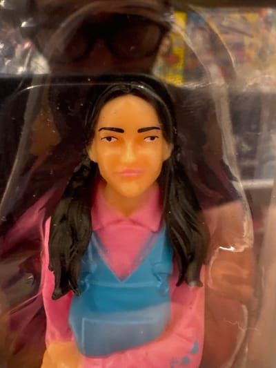 The media file shows a close-up, horizontal shot of a doll's head and upper body. The doll has fair skin, black hair styled into two pigtails, and painted facial features including large eyes with black lines for eyebrows and lashes, and pink lips. It appears to be wearing a pink and blue outfit. The doll is enclosed in clear plastic packaging, which has some smudges and reflections. The background behind the doll is blurred, but appears to show some colorful graphics, possibly from the toy's packaging. The lighting is bright, creating reflections on the plastic. The scene is likely a retail display or product packaging. There is no indication of time of day, weather, or specific location beyond the provided context of Keila, Estonia, and no discernible text is visible.