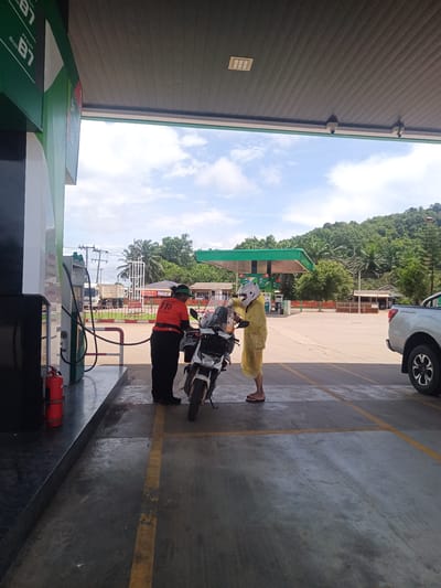 The photo captures a scene at a gas station in Plai Phraya, Thailand. An attendant in a green hat, black pants, and an orange shirt is interacting with a person wearing a white helmet and a yellow raincoat. The person is standing next to a white motorcycle. The gas station has green signage indicating fuel types. A truck is visible in the background. The sky is partly cloudy, and the surrounding area includes trees and a hill. The gas station is marked with yellow parking lines. A pickup truck is parked on the right side of the frame.