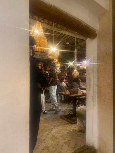 The image depicts an interior scene, possibly a restaurant or guesthouse, with a rustic ambiance. Visible elements include a wooden beam overhead, woven lampshades hanging from the ceiling, and a tiled floor. In the foreground, a person in a black garment stands partially obscured by a white wall. Further into the scene, several individuals are seated at tables, engaged in conversation or dining. One person is smoking. The lighting suggests it is evening or nighttime. The text "nza Guesthouse ANNEXE" is visible on a sign in the background. This establishment is located in Taghazout, Morocco.