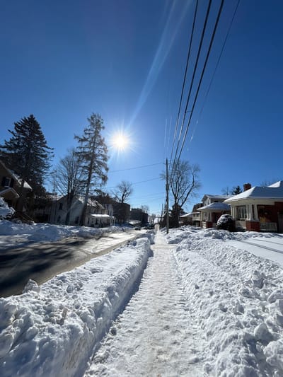 The image displays a snow-covered residential street in Kingston, Canada, under a clear, bright blue sky on a sunny winter day. The sun, a prominent starburst in the upper left quadrant, casts strong, defined shadows across the scene.

A paved two-lane street, partially cleared but with sections appearing wet and reflective, runs from the foreground into the distant vanishing point, where it appears to curve slightly downwards. Substantial, uneven snowbanks flank both sides of the street. Parallel to the street on the right, a cleared sidewalk extends into the distance, bordered by high snow accumulations. This sidewalk exhibits distinct tire tracks, suggesting recent snow removal by a small vehicle or snowblower.

Detached residential houses, primarily two-story structures with varying architectural styles, line both sides of the street. Their roofs are uniformly covered in a thick layer of white snow. On the immediate right, a house with a red brick facade and a prominent front porch with white columns is visible. Farther down the street, other houses with light-colored siding and snow-covered yards are discernible.

Bare deciduous trees, stripped of their leaves, are interspersed along the street, alongside several dark green coniferous trees, specifically pines or spruces. A series of utility poles, equipped with multiple overhead power lines, parallels the street on the right side, receding into the background. A single dark-colored vehicle is visible in the far distance on the street. No individuals or active interactions are present in the scene.