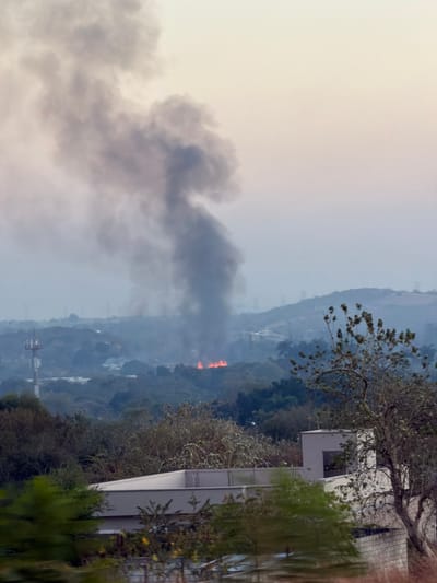 Gray smoke rises in a hazy landscape where a fire is visible in the distance. The fire appears as bright orange patches amidst the green trees. A modern building with visible balconies and trees is in the foreground, creating a contrast between the natural elements of the fire and the man-made structure. A hill stretches towards the sky in the background.