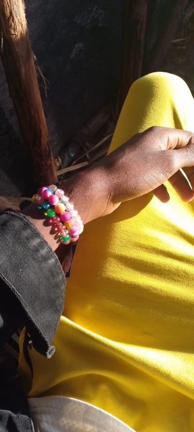 The image displays the lower left arm, hand, and a portion of the upper leg of an individual, observed within Matathani ward, Kenya. The individual exhibits dark skin tone, wears a black fabric garment on the left arm, and bright yellow fabric on the lower body, consistent with trousers or a skirt. The left wrist is adorned with multiple beaded bracelets, featuring an assortment of spherical beads in colors including pink, green, blue, yellow, and translucent white. Strong sunlight illuminates the subject from the upper left, creating distinct highlights and shadows. The background includes several rough-hewn wooden poles, a dark, textured fabric or mesh, and a partially visible dark object with a green circular component, possibly part of a vehicle, situated among dry vegetation elements.