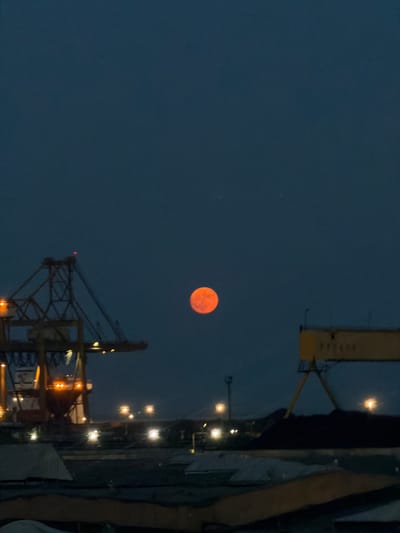 An orange-hued moon is visible in a dark blue twilight sky above an industrial port area. In the foreground, there are dark masses of material, possibly coal, and several structures that appear to be cargo or building materials. To the left, a large yellow and orange crane dominates the scene, with lights visible on its structure and at its base. To the right, a yellow gantry crane is partially visible. Several distant lights suggest activity within the port. The overall impression is one of industrial twilight in the Port of Constanța, Romania.