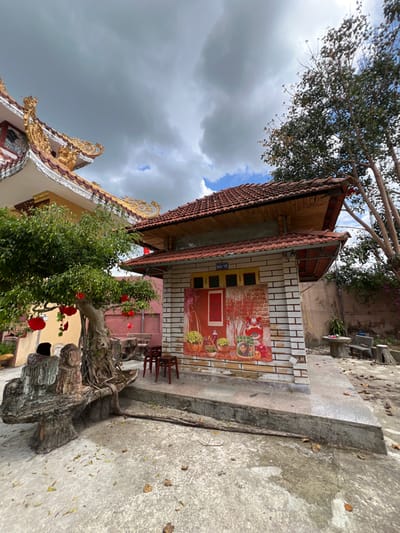 This image depicts an outdoor scene in Đăk Mar, Vietnam, featuring two distinct architectural structures, vegetation, and ground elements under a partially cloudy sky. On the left, a multi-story building with a light yellow facade features an ornate, tiered roof with golden-colored decorative elements, including dragon figures and curving eaves, typical of traditional Vietnamese temple architecture. To the right of this, a smaller, single-story building presents a red-tiled roof with visible wooden undersides. Its front wall is covered by a large, colorful mural or poster depicting a festive scene with red peach blossoms, potted kumquat trees, baskets of fruit, and a stylized figure, possibly associated with Lunar New Year celebrations. A red-framed door with a yellow trim is integrated into the mural. Above the mural, a sign reads "BÁO VỆ" and small yellow-framed windows are present.
In the foreground, a prominent bonsai-style tree with a thick, gnarled trunk and green foliage stands, adorned with several red lanterns. A large, sculpted concrete bench, designed to resemble natural rock, wraps around the base of this tree. Three dark wooden stools are positioned on a raised concrete platform in front of the mural-covered building. The immediate ground is a light-colored concrete surface, scattered with dry leaves. Towards the far right, a dirt patch contains a round concrete table and a matching concrete bench. Tall, slender trees with sparse foliage are visible on the right side of the frame, and lush green bushes partially obscure the base of the ornate building on the left. The sky shows a mix of heavy grey clouds and patches of clear blue. No individuals are present in the scene.