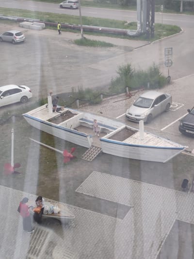 This image depicts an outdoor scene in Amursk, Russia, likely during the daytime with overcast weather. The foreground shows a large, weathered wooden boat that has been converted into a planter or decorative element. Two people, one in a red jacket and the other in dark clothing, are standing by another boat-shaped structure, which appears to be a bench. They are surrounded by several plastic bags, suggesting they may be returning from shopping.
In the middle ground, two children and an adult are interacting near the large wooden boat structure. One child is standing on the deck, while another, a young girl in a light-colored dress, is standing near a wooden ramp leading into the boat. An adult, dressed in dark clothing, is also present. Two large, red boat propellers are lying on the grass near the boat.
The background reveals a paved area with several parked cars, including a silver sedan and a dark SUV. A road with passing cars is visible further in the distance, with industrial-looking pipes and structures along the roadside. A person in a bright yellow vest is walking on a sidewalk near some concrete blocks and pipes. The overall impression is a somewhat industrial or utility area adjacent to a residential or public space, with elements of urban life and leisure.