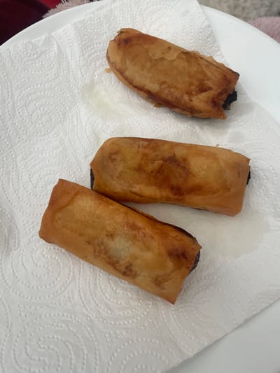 Three golden-brown spring rolls are arranged on a white paper towel. The paper towel has a raised, circular pattern. The spring rolls appear to be deep-fried and are slightly uneven in shape. One spring roll is positioned at the top of the frame, slightly tilted. Two other spring rolls are stacked below it, one directly beneath the other, oriented horizontally. The background is blurred and appears to be a white plate and a hint of a red and white striped fabric. There is no indication of the location in Bendigo, Australia.