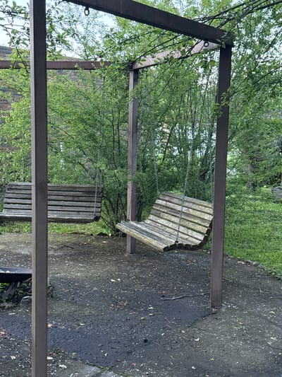 The image depicts a two-seat wooden swing set structure under a canopy of green leaves. The swing set is made of dark metal posts and beams, supporting two weathered wooden swings. One swing is positioned to the left, parallel to the ground, and appears empty. The other swing, to the right, is tilted downwards as if recently occupied, and also appears empty. The ground beneath the swing set is a dark, wet surface, possibly asphalt or packed dirt, with scattered leaves and debris. In the background, a brick building is partially visible through dense green foliage. The scene appears to be in an outdoor setting, likely a garden or park area within the city of Redant, Russia. There are no people visible in the image.