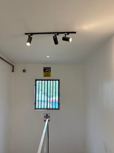 The image depicts an interior scene with white walls and a white ceiling. A black track light with three adjustable spotlights is mounted on the ceiling. To the left, a red pipe runs diagonally along the wall. There is a small, circular security camera mounted on the wall. Below the camera, a square window with black bars is visible, offering a view of trees and some buildings outside.
A yellow and black "NOTICE" sign is posted on the wall above the window, with text that reads "PLEASE TURN OFF ALL ELECTRICAL APPLIANCES." Below the window, a circular sign with a person using a handrail is visible, indicating "Use handrail." To the right of this, a red and white sign with text that is partially obscured reads "DO NOT". A white railing for a staircase is partially visible in the foreground, extending upwards from the bottom of the image. The overall environment appears to be a clean, indoor space, possibly a hallway or stairwell. The location is identified as Gwaska, Nigeria.