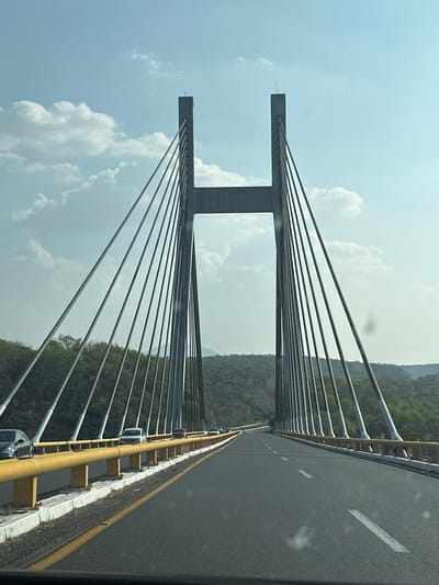 A daytime, eye-level shot from a moving vehicle, looking up at a cable-stayed bridge, presumed to be located in Tlanipatla, Mexico. The bridge is the primary subject, with two large, gray towers perpendicular to the road, supporting numerous suspension cables that converge towards the roadway. The cables are silver-gray. The road surface appears to be new asphalt, marked with lane dividers. The sides of the bridge have a yellow guardrail over a white concrete base. The background shows a forested area on a hill. The sky is partly cloudy. There are no people visible.