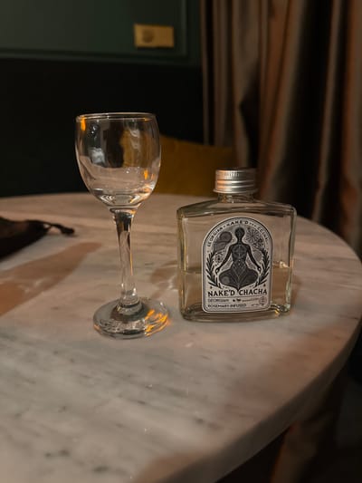 The image shows a clear glass tasting shot glass and a small, clear glass bottle of NAKE'D CHACHA Georgian Rosemary-Infused spirit on a white marble tabletop. The bottle has a silver metal cap and a label featuring a black and white illustration of a person meditating within a circular design, surrounded by text that includes "GEORGIAN NAKE'D CHACHA" and "ROSEMARY-INFUSED". The background is dark and out of focus, with draped brown curtains and a hint of a dark green wall with a gold-colored fixture. The setting is likely an indoor establishment such as a bar or restaurant, possibly in Tsinandali, Georgia, a region known for its wine and spirits production. There are no people visible in the image.