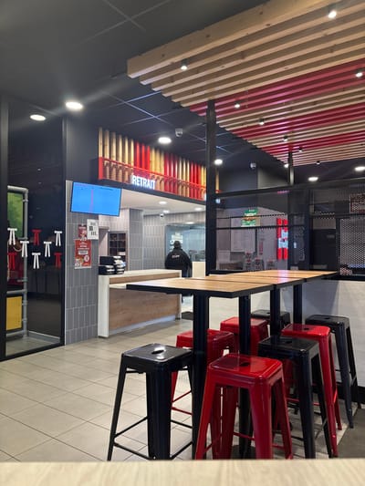 The image displays the interior of a modern fast-food restaurant in Fegersheim, France. The foreground shows a seating area with multiple tall, square tables topped with light-colored wood, supported by black metal bases. Around these tables are numerous glossy bar stools, predominantly red and black, with a few others of indeterminate color visible further back. The floor is covered in light grey tiles.

In the midground, a service counter features a light wood front and a white countertop. Behind this counter, one individual, identifiable as "STAFF" by text on their dark jacket, stands facing away from the camera. Above the counter, a red illuminated sign spells "RETRAIT" (pickup) in capital letters, positioned beneath a decorative paneling of alternating vertical wooden and red slats. To the left of the "RETRAIT" sign, a blue digital screen displays order statuses, including "PREPARING". The wall behind the counter is clad in light grey vertical tiles, with some small promotional flyers attached.

To the far left, a children's play area with green, yellow, and grey piping and white "T"-shaped connectors is partially visible through a glass partition. To the right, a black perforated metal screen acts as a room divider. The ceiling features a distinctive design with alternating light wooden and red painted beams, interspersed with recessed lighting fixtures and several visible security cameras. The overall environment is brightly lit and appears clean, with no direct customer interactions depicted.