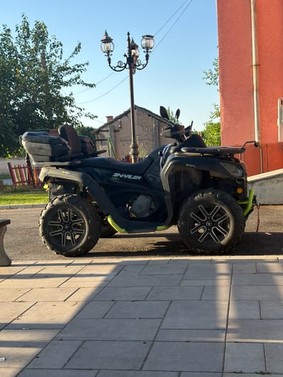 The image shows a black and green quad bike parked on a paved area next to a building. The quad bike has the brand name "SNARLER AT6 L" visible on its side. It is a sunny day, and the light suggests it is late afternoon, with long shadows cast across the pavement. The setting appears to be a residential or semi-rural area, with a building with a reddish-pink wall on the left and trees and greenery in the background. A vintage-style street lamp with two globes is also visible. The quad bike is the primary subject, and it is stationary. There are no people in the image. The overall mood is calm and still, capturing a snapshot of the vehicle in its environment.