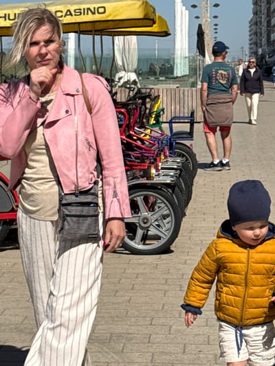The image depicts an outdoor scene on a sunny day, likely a promenade or boardwalk in a coastal town, with people and recreational vehicles. In the foreground, a woman with blonde hair, wearing a pink leather jacket, a light-colored top, and striped wide-leg pants, smiles slightly while looking towards the camera. Part of a yellow canopy structure next to her displays the text "CASINO" and a partial "HU".
Adjacent to the woman is a row of brightly colored rental bicycles or quadricycles, equipped with large wheels, parked on a wooden platform. These vehicles appear to be designed for leisure.
Further in the mid-ground, a man wearing a blue baseball cap, a dark green t-shirt with an abstract graphic, and brown shorts, stands facing away from the camera, looking towards the right. In the distance behind him, two other individuals are visible.
In the upper right portion of the frame, a young child wears a dark blue beanie, a yellow puffer jacket, and light-colored striped shorts, looking upwards with an outstretched arm.
The ground is a light-colored paved surface, and tall, modern buildings are visible in the distant background under a clear blue sky. The strong shadows indicate bright sunshine, suggesting midday or early afternoon. A small, grey fanny pack worn by the woman has the faint text "SPORTS" on a label. The overall atmosphere is recreational and pleasant. The "CASINO" sign and rental bikes are strong location cues for a tourist area such as Middelkerke, Belgium.
