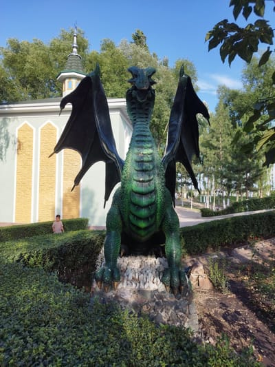 In the city of Kenbulun, Kyrgyzstan, a large, green dragon sculpture stands prominently in an outdoor setting. The sculpture depicts a mythical creature with outstretched wings, sharp claws, and a detailed, scaly texture. It is positioned on a rocky base surrounded by lush green hedges. Behind the dragon, a light-colored building with decorative yellow and white accents is visible, topped with a distinct green-roofed tower that has a pointed spire. A young boy, dressed in a light brown shirt, is standing amidst the hedges to the left of the sculpture, looking towards the building. The environment appears to be a park or garden, with trees and blue sky forming the backdrop. The scene is captured during daylight, with sunlight casting shadows on the ground.