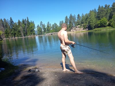 This image depicts a person fishing on the shore of a body of water. The subject is a man, shirtless and wearing patterned swim trunks, who is holding a fishing rod and appears to be casting or reeling in. He is standing on a rocky shoreline, with a pair of black and white striped flip-flops nearby. The water is calm and reflects the clear blue sky and the surrounding coniferous trees. In the background, a wooden bridge can be seen spanning the water, and more trees line the opposite bank. There are also other people visible in the distance, some appearing to be swimming or relaxing by the water. The scene suggests a warm, sunny day, likely in the summer, given the attire of the people and the lush greenery. The overall mood is relaxed and recreational. The location is identified as Drammen, Norway. No visible text is present in the image.