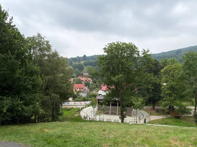 This is a photograph of an outdoor scene in Świeradów-Zdrój, Poland. The foreground is a grassy slope. In the middle ground, there is a paved area with benches and a covered pavilion with railings. To the left of the paved area, there is a fence and a road. Further in the distance, there are several houses with red roofs nestled among trees. The background consists of a wooded hillside under a cloudy sky.