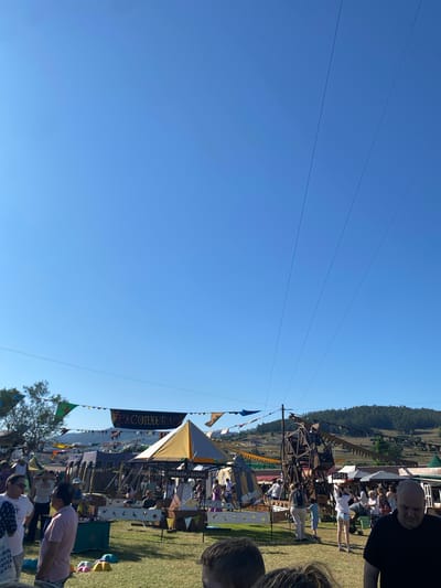 The image captures an outdoor event, possibly a fair or festival, under a clear blue sky. In the background, a lush, tree-covered hill forms the horizon. The foreground is occupied by a grassy field where various stalls and attractions are set up. Several people are visible, some interacting with each other and others browsing the stalls. A large banner with "EXCONXURAC" is prominently displayed, suggesting the name of the event. There is a prominent wooden structure resembling a wheel or carousel, and other tents and marquees are spread across the field. The atmosphere appears festive and lively, indicating a public gathering on a sunny day. This event is likely taking place in the city of Llanera, Spain, as indicated by the context.