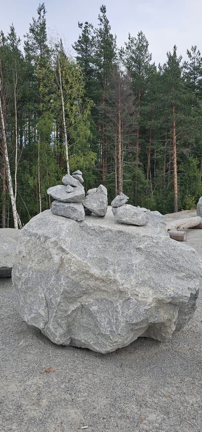 A large, gray, granite boulder is prominently displayed in the foreground. Several smaller, gray stones are balanced on top of the large boulder, forming a rock cairn. In the background, a dense forest of green pine and birch trees stretches across the horizon. The ground is covered with small gray gravel. The scene appears to be an outdoor setting, possibly a park or natural area.