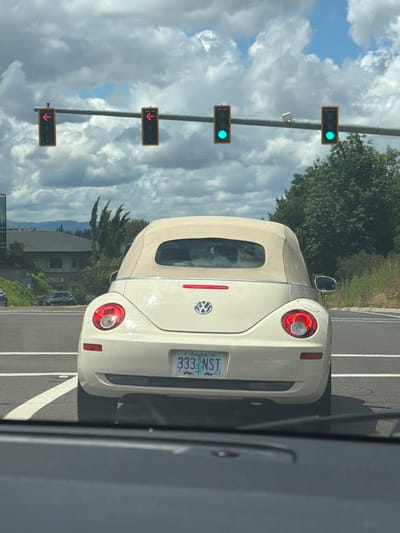 A white convertible Volkswagen Beetle is viewed from behind, driving on a road. The car has its top down, and a red brake light is illuminated. The license plate reads "333 NST" and is from Oregon. The scene appears to be in Beaverton, USA, based on the provided context.
In the foreground, to the left of the car, a series of traffic lights are visible. The top two lights are green, while the bottom two lights display red downward arrows. The sky is partly cloudy with a mix of blue sky and white clouds. Trees and foliage are visible on the right side of the road and in the background, suggesting it is daytime with mild weather. The overall impression is that the car is stopped at a traffic signal. A portion of another vehicle, appearing to be a dark gray SUV, is visible on the right edge of the frame, indicating the perspective is from inside another vehicle.
