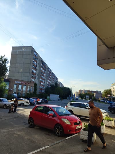 In Zhukovsky, Russia, on a clear day with a blue sky and scattered clouds, a street scene unfolds. A large, grey, multi-story apartment building dominates the background, its facade showing rows of windows and balconies. In front of it, a smaller brick building houses a medical clinic, identified by signage.
The foreground features a paved street and sidewalk area. Several vehicles are parked along the street. A red hatchback is prominently positioned in the lower right quadrant of the image. To its left, a white SUV is parked, and further back, a yellow taxi with visible damage to its rear bumper is also parked. A woman is seen with a child in a stroller near the taxi.
A man in a brown polo shirt and black pants is walking in the foreground, heading towards the right side of the frame. He is wearing dark sandals. The environment includes streetlights and some greenery. The overall impression is that of a typical urban residential and commercial area in the afternoon.