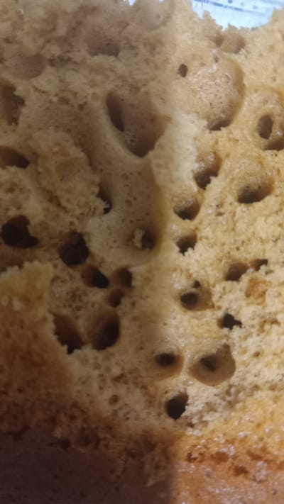 The image is a close-up, top-down view of the porous, golden-brown interior of a baked good, possibly a cake or bread. The texture is irregular, with numerous air pockets and small holes of varying sizes and depths. The lighting suggests an indoor environment, and the background, visible at the top edge, appears to be a light blue and white striped pattern, possibly a plate or tablecloth. There is no indication of a specific location within Oliveira do Bairro, Portugal, as the image focuses solely on the texture of the food item.