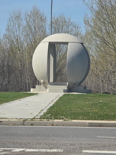 The image depicts an outdoor scene in Polígono Industrial Nicomedes García, Spain. Centrally positioned is a large, light-grey spherical sculpture, likely constructed from stone or concrete, featuring a distinct vertical, U-shaped opening or void on its front face. The sculpture rests on a multi-tiered, stepped base of matching light-grey material. A straight, light-grey paved path leads towards the base of the sculpture, flanked by strips of green grass containing scattered small white daisy-like flowers and a few yellow dandelions. In the immediate foreground, an asphalt road with a single white painted line runs horizontally, separated from the grass by a light-grey concrete curb. The background consists of a dense line of deciduous trees, mostly bare or exhibiting very sparse, light-green early spring foliage, set against a clear, light blue sky. A thin, dark vertical pole extends upwards behind the right side of the sculpture. No individuals or active subjects are present.